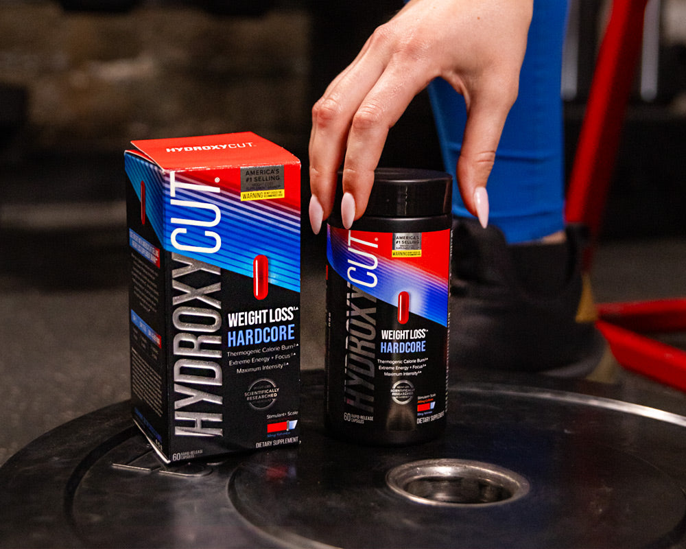 Hydroxycut original bottle on a counter with a glass of water and a woman holding two capsules